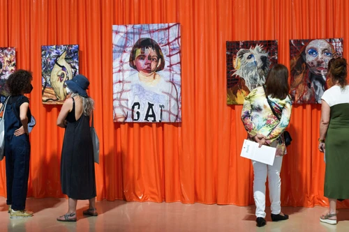 Besucherinnen betrachten farbintensive Porträtgemälde vor leuchtend orangefarbenem Hintergrund in einer Galerie.