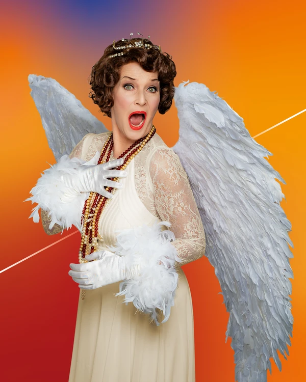 A person in a white angel costume with large feathered wings and a crown poses expressively against a vivid background.