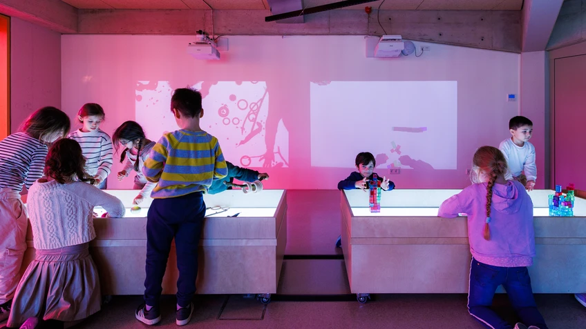 Eine Gruppe von Kindern spielt in einem abgedunkelten Raum an leuchtenden Tischen. Einige bauen mit bunten Bausteinen, andere experimentieren gemeinsam an einem Lichttisch. An der Wand werden farbige Projektionen sichtbar, die von den Aktivitäten der Kinder stammen. Die Atmosphäre wirkt lebendig und spielerisch.