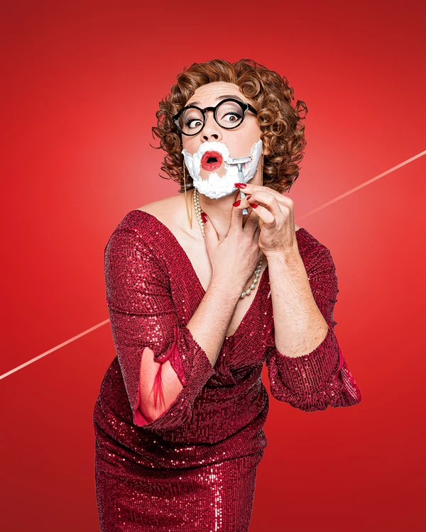 A person in a sparkly red dress with pearls and curls shaves their face against a red background.