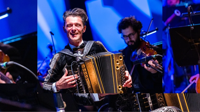 Konzertfoto von Herbert Pixner mit einer Ziehharmonika, hinter ihm zu sehen: das Tonkünstler Orchester Niederösterreich auf einer blau beleuchteten Bühne.