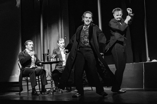 Bühnenszene "Die Fledermaus": an einem Tisch sitzen Gabriel von Eisenstein (Raphael von Bargen), Dr. Falke  (Peter Lesiak) im Vordergund tanzen:  Prinz Orlofsky (Moritz Mausser), Frosch (Sebastian Wendelin);