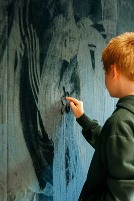 Ein Junge zeichnet mit seinem Finger Muster auf eine mit dunkelgrünem Samtstoff überzogene Wand.