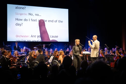 Foto von "Orchesterkaraoke", auf der Bühne ein Orchester, dahinter wird offenbar ein Songtext projiziert, eine Person steht an einem Mikrofon und singt.