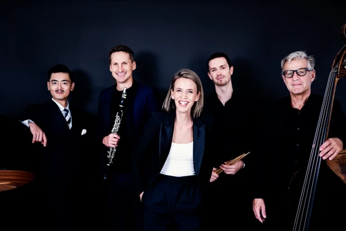 5 Musiker:innen, dunkel gekleidet, vor dunklem Hintergrund