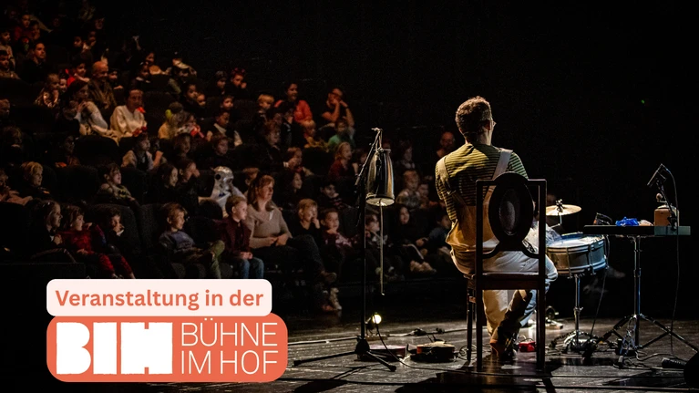 Im Vordergrund ist der Multiinstrumentalist der ZONZO Compagnie zu sehen, hinter ihm, ihm zugewandt, ist ein Publikum zu sehen.