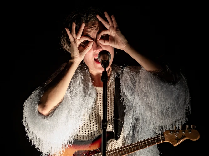 Photo of Aline Goffin, her fingers forming glasses in front of her eyes, an electric guitar hanging from a strap in front of her, wearing a striking glittery fringed jacket. 