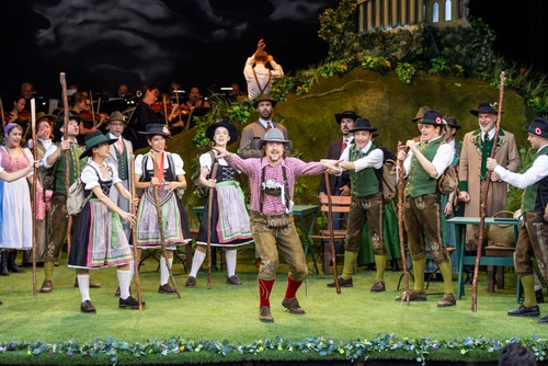 A wide-angle shot of a large ensemble scene on a stage. Numerous people in traditional costume are spread across the entire width of the stage. Many are wearing hats, waistcoats, long skirts or shorts with stockings. Some are holding baskets, whilst others have their arms raised or outstretched as part of a choreographed movement. In the background, additional figures are visible on raised platforms or steps. The backdrop depicts a rural setting with green fields and a dark sky. In the centre of the foreground, a person wearing a red-and-white chequered shirt, leather trousers and a hat strikes a dance pose, arms outstretched. The entire scene appears densely populated and dynamically lit.