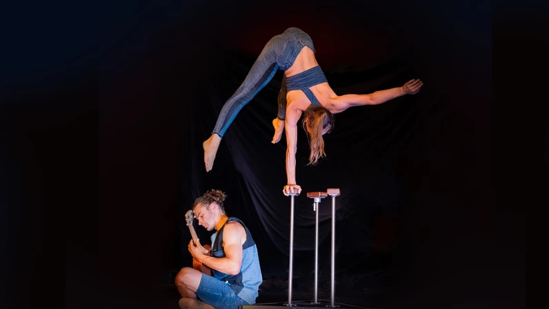 Eine Artistin steht in einem einarmigen Handstand auf erhöhten Handstandstützen, während neben ihr ein Musiker im Sitzen Gitarre spielt, beide in sportlicher Kleidung vor dunklem Hintergrund.