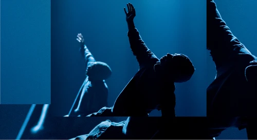 Tänzer:innen der Compagnie Far From The Norm performen "BLKDOG" von Choreograf Botis Seva. Auf einer spärlich in blau beleuchteten Bühne vor schwarzem Hintergrund performen zwei Tänzer:innen eine Figur, einen Arm in die Höhe gestreckt. 