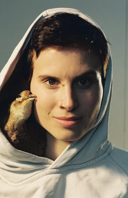 Nahaufnahme einer Frau mit dunklen kurzen Haaren, heller Hoodie mit Kapuze auf dem Kopf, ein Vogel schmiegt sich an ihr Gesicht