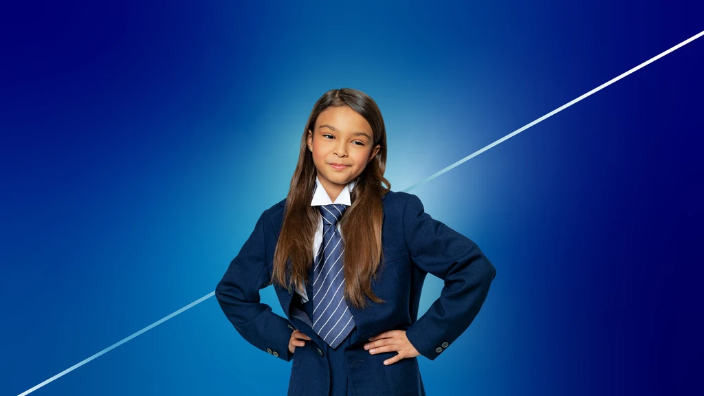 Ein circa neunjähriges, braunhaariges Mädchen in einer dunkelblauen Schuluniform mit Krawatte steht mit Händen in die Hüften gestützt vor einem blauen Hintergrund. Nahaufnahme.