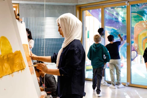 In the foreground, a girl paints at an easel, while in the background, three boys look through a glass door at a colourful textile sculpture by Toshiko Horiuchi MacAdam.