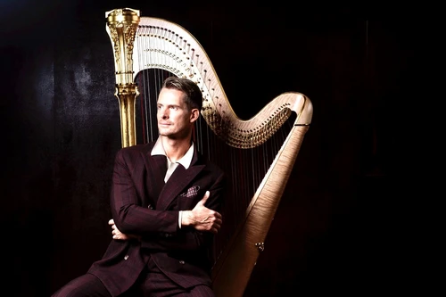 Foto von Harfinist Xavier de Maistre, elegant gekleidet in einem dunklen Raum vor schwarzem Hintergrund. Direkt hinter ihm steht sein Instrument, die Harfe, gegen die er sich leicht anlehnt.