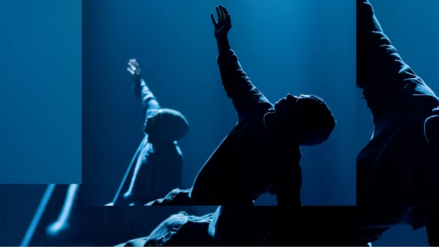 Tänzer:innen der Compagnie Far From The Norm performen "BLKDOG" von Choreograf Botis Seva. Auf einer spärlich in blau beleuchteten Bühne vor schwarzem Hintergrund performen zwei Tänzer:innen eine Figur, einen Arm in die Höhe gestreckt. 