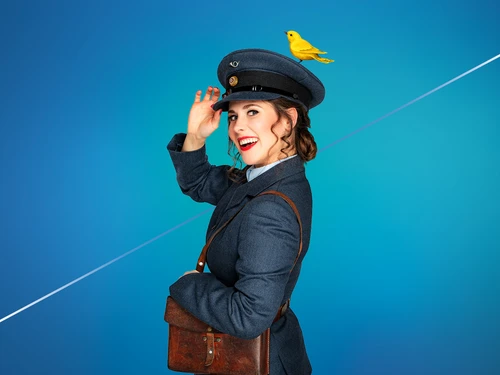 Eine Person in historischer Postuniform mit blauer Mütze und Mantel lächelt und tippt die Mütze an, während ein gelber Vogel darauf sitzt.