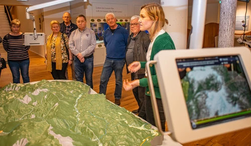 Im Vordergrund ist ein Tablet zu sehen, das eine Maske auf das Landschaftsmodell projiziert, anhand der man die Entwicklung des Waldes nach der letzten Eiszeit beobachten kann. Im Hintergrund ist ein Guide und Besucher*innen um das Landschaftsmodell versammelt. 