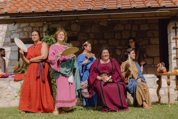 Roman matrons in colorful tunics stand and sit in front of the portico and look at the camera. 