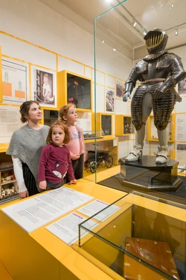 Frau mit 2 Kindern blicken auf Kinderharnisch in einer Vitrine.
