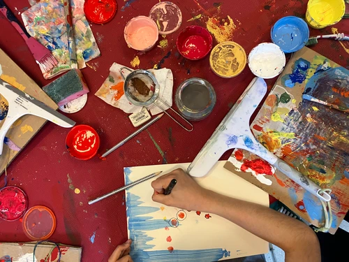 Blick von oben auf eine kreative Arbeitsszene mit Farben, Pinseln und einer Person, die auf Papier malt.