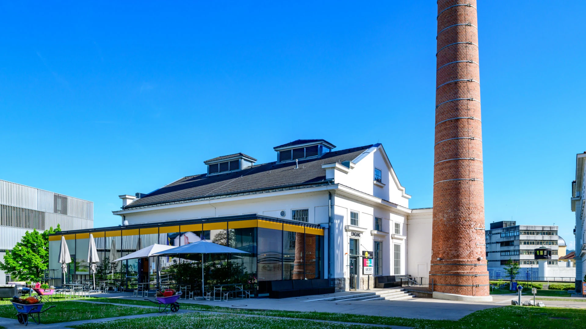 Historisches Industriegebäude mit hohem Ziegelkamin und angeschlossenem Cafébereich mit Sonnenschirmen.