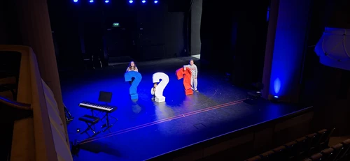 Drei Kinder mit drei großen Fragezeichen auf einer dunkelblau beleuchteten Bühne