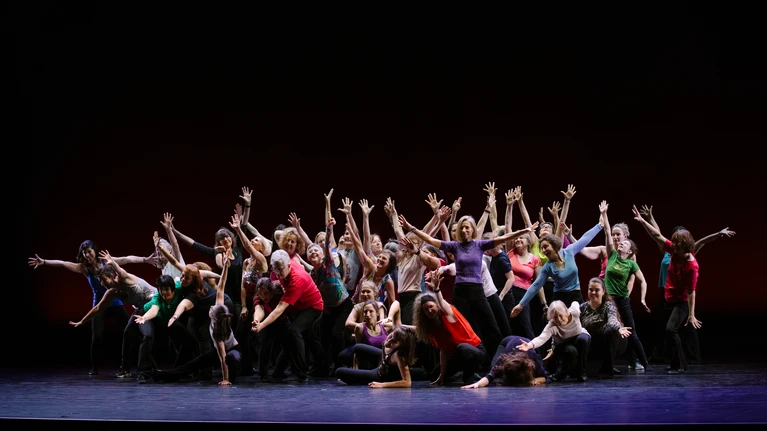 Foto von der Winter-Werkschau 2025 mit einer der Tanz-Communities vom Festspielhaus St. Pölten, bunt gekleidet in einer Gruppenpose auf einer schwarzen Bühne.