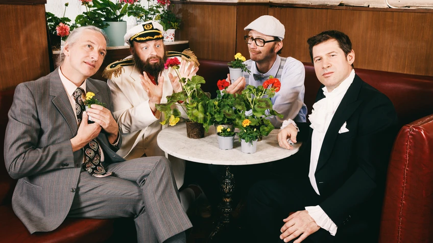 Vier Männer sitzen um einen Kaffeehaustisch auf einer roten Lederbank, auf dem Tisch stehen Blumen in Töpfen. Ein Mann trägt ein Kapitänskostüm
