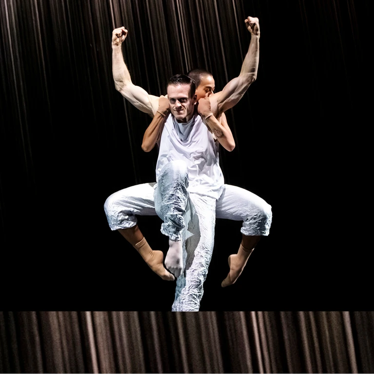 Zwei Tänzer:innen performen "Mémoires d’Oubliettes", eine Choreografie von Jirí Kylián. Ein Tänzer in weißem Kostum steht im Vordergrund auf einem Bein, das andere vor sich angewinkelt, seine Arme seitlich nach oben, die Armmuskeln stark angespannt. Hinter ihm ist eine weitere Person zu sehen, ebenfalls in weißem Kostüm, die sich an seinem Rücken festklammert, beide Beine angehoben und seitlich angewinkelt.