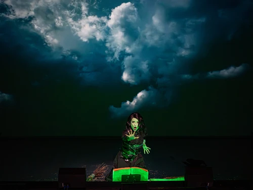 Black-haired woman with green skin in a dramatic pose, hands stretched forward, a green-lit open book in front of her, dark, dramatic clouds in the background