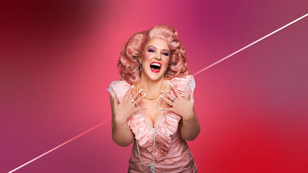 Nahaufname einer Frau in rosa Rüschenkleid, langer Perlenkette, rosa Perücke die vor einem rosafarbenen Hintergrund zur Kamera nach vorne gebeugt steht. Sie lacht mit weitgeöffnetem Mund.