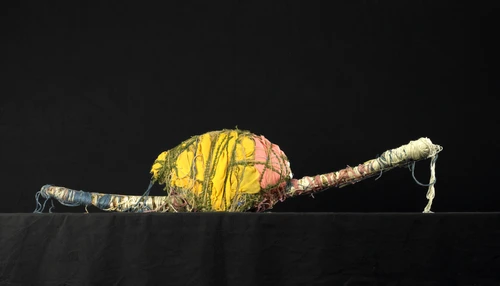 Längliche textile Skulptur aus eng umwickelten Fäden und Stoffen in Gelb, Rosa, Grün und Blau, mit einer zentralen, kugelförmigen Verdichtung auf schwarzem Hintergrund