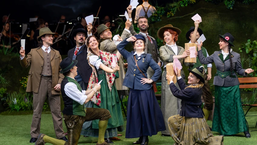 A stage photograph featuring a large group in traditional-looking costumes. The people are standing close together on a stage decorated in green. Several figures are wearing hats, waistcoats and long skirts, and are holding letters and other documents aloft whilst laughing. In the centre is a person in postal service uniform, who, whilst laughing, is also dutifully holding their right hand to their cap. In the background is a natural-looking backdrop featuring plants and a dark sky illuminated by blue lighting. The lighting highlights the entire group evenly.