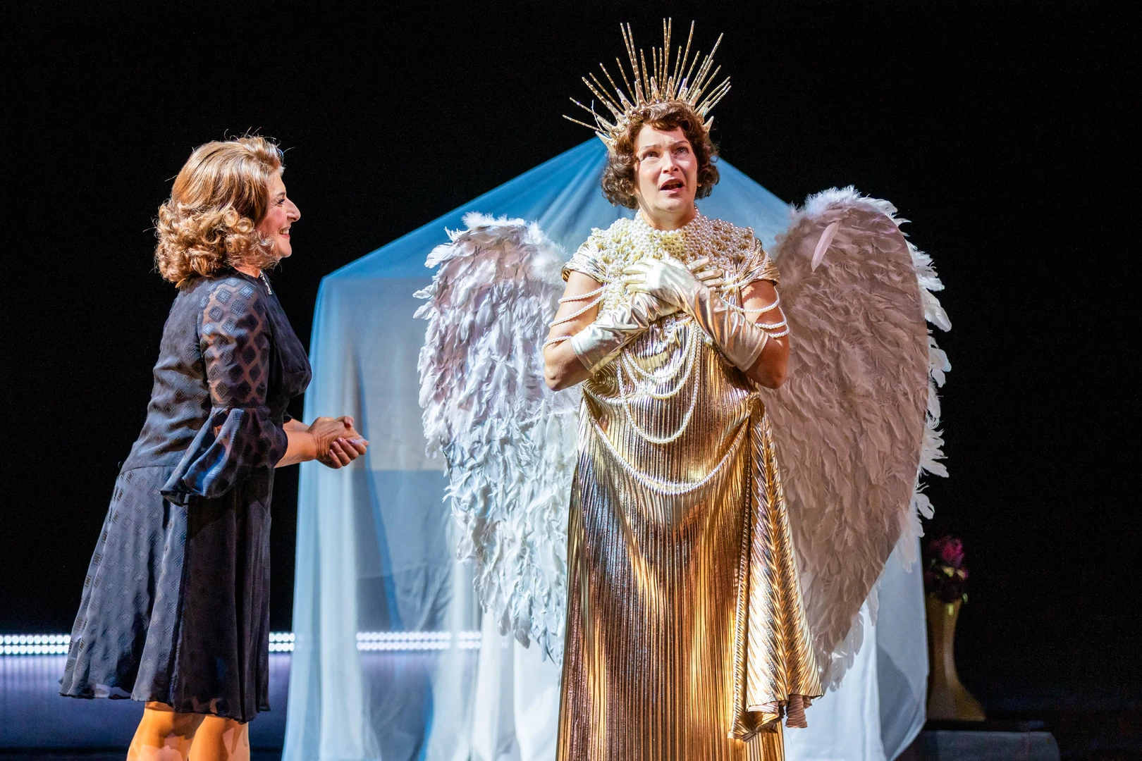 Two people are standing side by side on a theatre stage. One is wearing dark clothes, the other a gold-coloured costume with wings and a radiant headdress. In the background is a piece of furniture covered with a white, transparent cloth.