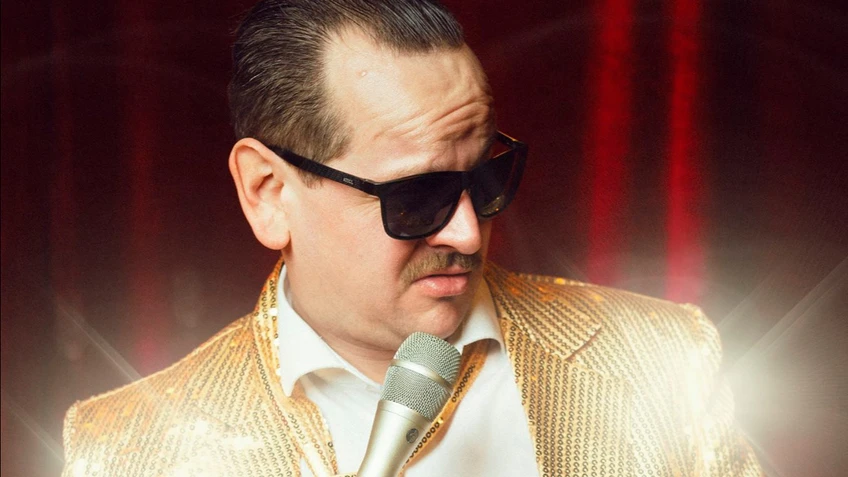 Romantic Slivo steht mit dunkler Sonnenbrille und Schnurrbart vor einem roten Bühnenvorhang, trägt einen auffälligen goldenen Glitzeranzug und hält ein Mikrofon mit Kabel in der Hand, während er seitlich nach unten blickt.