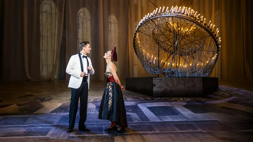 Zwei Darsteller stehen auf einer Bühne vor einem großen, auf dem Boden liegenden kunstvollen Kronleuchter mit vielen Kerzen. Die Person links trägt einen weißen Smoking mit schwarzer Fliege, die Person rechts ein elegantes schwarzes Kleid mit rotem Oberteil und Kopfschmuck. Sie diskutieren miteinander. Der Hintergrund zeigt hohe Fenster und warme Beleuchtung.