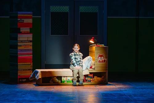 A child sits on a bench on a small stage, surrounded by a tall stack of colourful books and a round table with a lamp. The scene is warmly lit, and the background shows closed doors in dark colours.