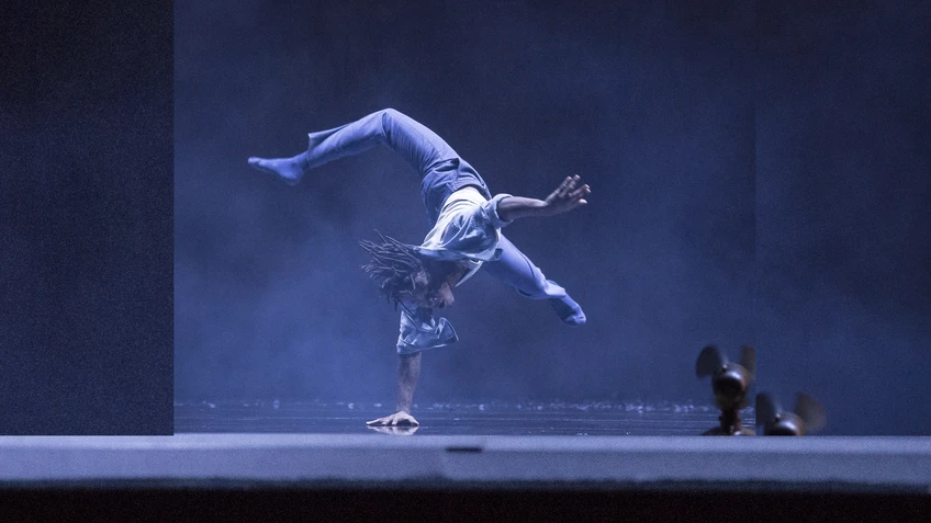 Performancefoto aus dem Tanzstück "Kaléidoscope", choreografiert von Mourad Merzouki, getanzt von der Compagnie Käfig. Ein Tänzer tanzt eine akrobatische Figur, bei der er lediglich mit einer Hand den Boden berührt, der Rest seines Körpers ist in der Luft, ein Bei nach hinten im Sprung abgewinkelt. Die Bühne ist in blaues Licht und durchsichtigen Bühnennebel gehüllt.