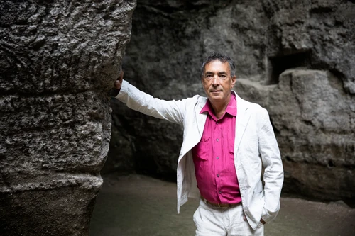 Foto von Hubert von Goisern, er trägt ein rosa Hemd und weiße Anzugjacke und Anzughose. Mit einer Hand lehnt er an eine Steinwand, auch im Hintergrund ist eine Steinwand zu sehen.