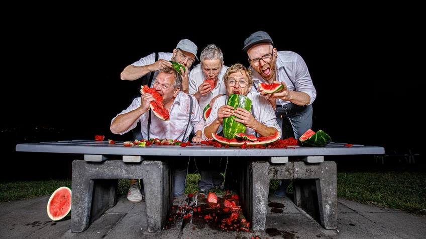 Die fünf Achterl beugen sich in weißen Hemden lachend und wild essend über eine Parkbank voller zerschmetterter Wassermelonen, während sie große Stücke der Frucht in den Händen halten und die Szene humorvoll chaotisch wirkt.