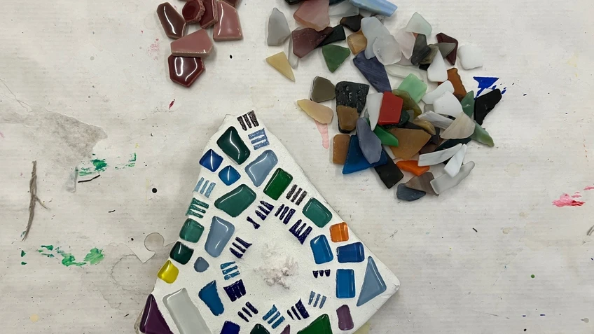 Dreieckiges Mosaik aus bunten Glasstücken auf einem Tisch, daneben lose Scherben in verschiedenen Farben und Formen.