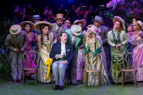 A group scene featuring several people in colourful, period-style costumes. The group is standing close together on a stage; some of the figures are wearing hats and holding small props. Their clothing features bold colours such as purple, green and yellow, as well as patterned fabrics. They are all facing a singer in the foreground, who is the only one seated in an armchair. The background is dark and filled with stage elements and other figures. The lighting clearly highlights the group in the foreground and makes the colours of the costumes glow.