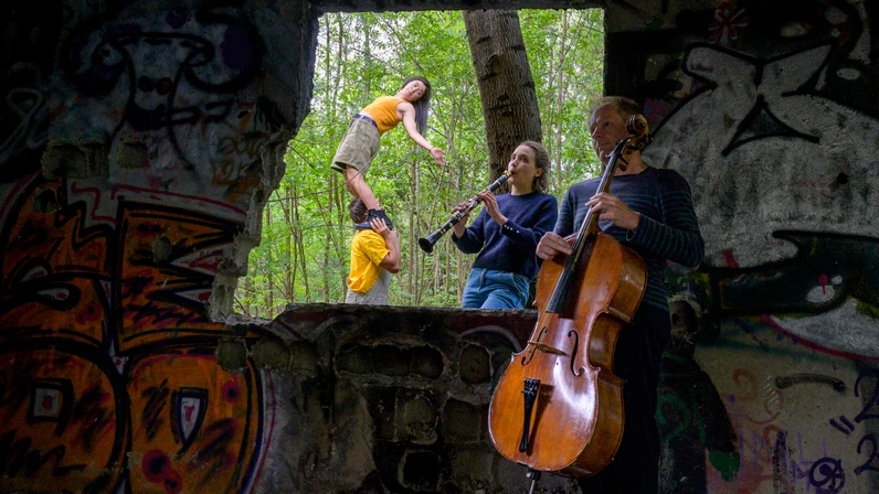 In einer verlassenen, bunt besprühten Ruine spielen ein Cellist und eine Klarinettistin Musik, während im Hintergrund eine Akrobatin auf den Schultern eines Partners balanciert und durch ein großes Fenster der grüne Wald sichtbar wird.