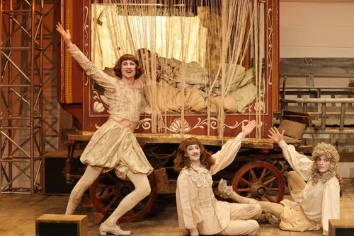 Menschen alten beigen Kostümen und Perücken strecken einen Arm in die Höhe, dahinter ein alter Wagen mit Vorhang, hinter dem eine Person in Kissen und Decken liegt.