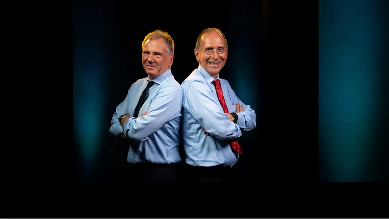 Foto von Journalist und ORF-Moderator Armin Wolf und der ebenfalls durch den ORF bekannte Politikwissenschaftler Peter Filzmaier stehen – beide mit hellblauem Hemd und Krawatte – Rücken an Rücken aneinander vor einem schwarzen Bildhintergrund und lächeln seitlich zur Kamera.