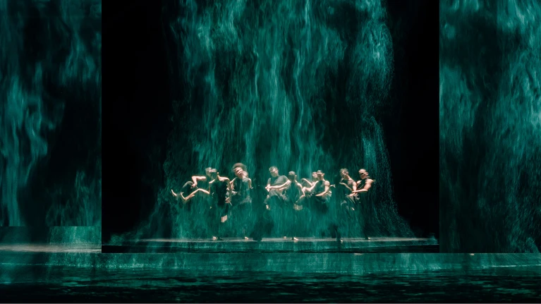 Stage photo from ‘All Ears’, a performance from the Cloud Gate Dance Theatre of Taiwan by choreographer Cheng Tsung-lung. The dancers, dressed in black, are arranged in a circle, holding hands and jumping.