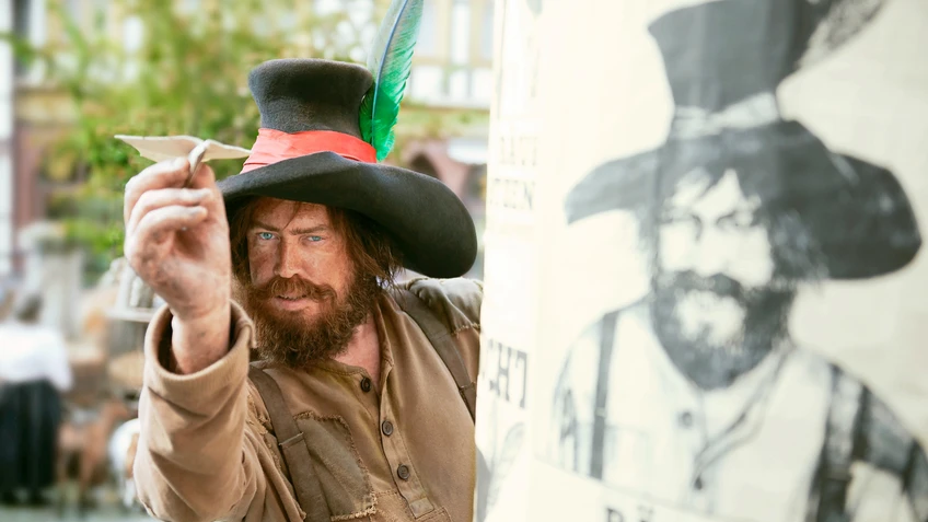 Ein Mann in geflicktem braunem Hemd mit großem schwarzen Hut mit grüner Feder hält einen Papierflieger, er ist schmutzig im Gesicht. Daneben eine Säule mit seinem Portrait gezeichnet