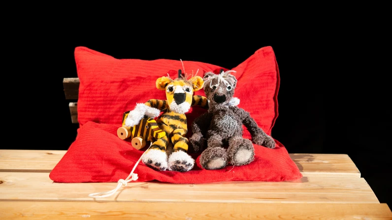 Zwei weiche Tierpuppen – ein Tiger mit Tigerente an der Hand und ein Bär – sitzen eng nebeneinander auf einem roten Kissen auf einem Holztisch vor schwarzem Hintergrund.
