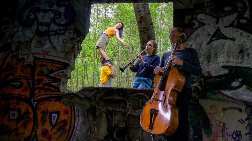 In einer verlassenen, bunt besprühten Ruine spielen ein Cellist und eine Klarinettistin Musik, während im Hintergrund eine Akrobatin auf den Schultern eines Partners balanciert und durch ein großes Fenster der grüne Wald sichtbar wird.