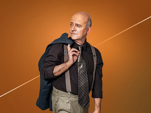 Ein Mann mittleren Alters steht vor einem orangefarbenen Hintergrund, trägt Hemd, Krawatte und Hosenträger, mit einem blauen Sakko über der Schulter.